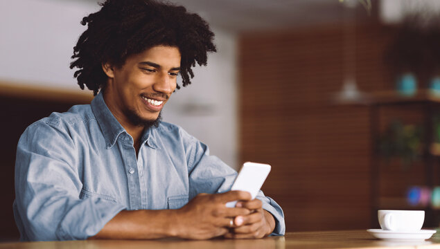Smiling millennial attractive african american curly man typing on smartphone at table with coffee in cafe interior, free space. Blog, great offer, app, chat in social media, modern communication - Powered by Adobe