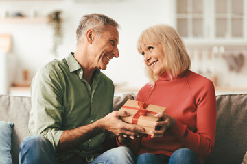 Senior Husband Giving Gift Box To Wife Congratulating Her On Birthday Or Anniversary Sitting On Couch At Home. Mature Couple Celebrating Family Holiday Together Indoors. Romantic Present Concept