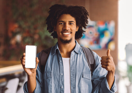 Young African American Guy Holding Smartphone With Blank White Screen And Gesturing Thumb Up While Standing Indoors In Cafe, Happy Smiling Black Man Recommending New App Or Website, Mockup Image