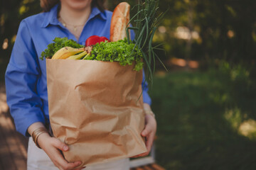 woman carries brown paper bag with fresh vegetables and colorful fruits in lush garden, enjoying...