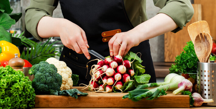 In bright kitchen, private chef chops fresh radishes and colorful vegetables, herbs onwooden cutting board