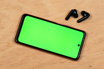 a wireless earphones with a smart phone with a green display screen over a wooden background