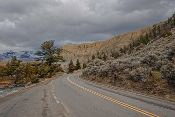 A winding road follows the Yellowstone river, surrounded by rugged mountains and trees. Dark clouds loom overhead, hinting at approaching weather during a peaceful rural day.