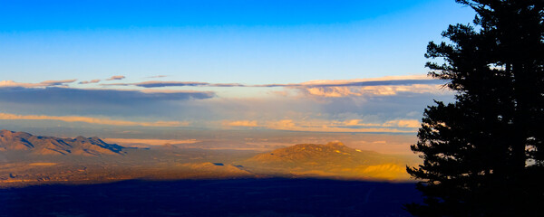 Fototapeta premium A breathtaking sunset paints the sky with warm colors as the sun sets behind the mountains. Looking west from Sandia Peak Albuquerque New Mexico. Trees stand tall, silhouetted against the vibrant hor