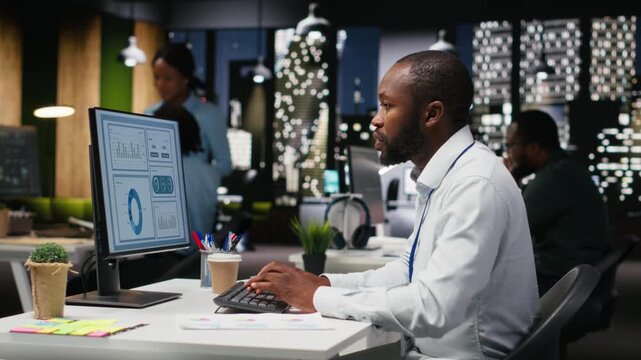 Black business consultant proofreading financial numbers at desktop after hours, studying data for monthly performance reports. Ensuring company objectives achievement at night. Camera B.