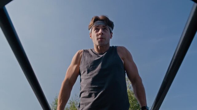 Young male athlete wearing grey t-shirt and headband performing dips on parallel bars of sports facilities, then stopping doing exercise and having short break