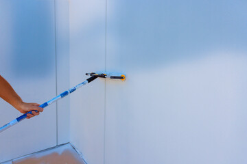 Worker applies white paint to wall with roller brush in well lit space, renovation work in progress.