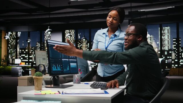 African american workers team consulting each other in solving tasks at night, completing monthly reports and reviewing KPI data insights for productivity. Collaboration for success. Camera A.