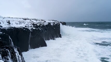 Powerful ocean waves crash against snow-covered volcanic cliffs under a dramatic sky. Wild winter coastal landscape with rugged beauty and raw force. - Powered by Adobe