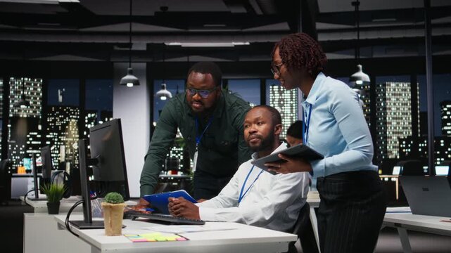 African american staff members consulting each other for monthly reports, reviewing KPI data insights for business productivity late at night. Collaboration between employees for success. Camera A.
