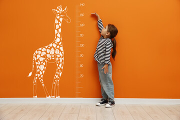 Fototapeta na wymiar Cute little girl measuring her height near orange wall with scale and drawing of giraffe