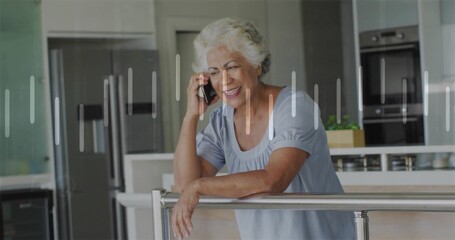 Leaning senior woman holding smartphone to ear in modern kitchen, with railing and potted plant