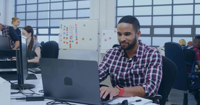 Typing man in red-and-white checked shirt at desk in open-plan office, with laptop and whiteboard
