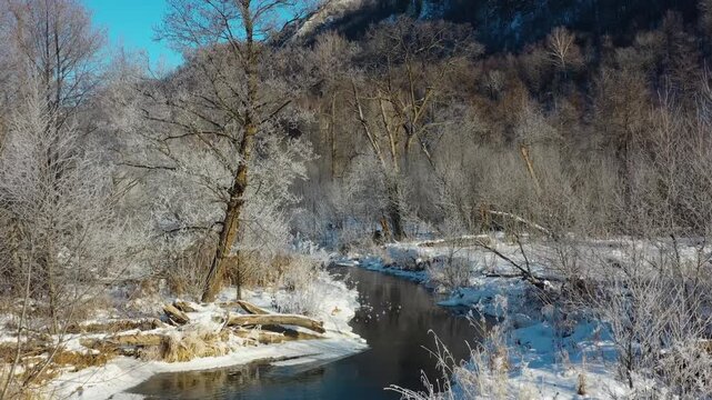 A scenic view of a tranquil river flowing through a snow covered forest in the South Urals, Russia. The winter sun illuminates the frosted trees and the distant mountain range, creating a peaceful and
