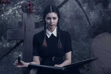 A young woman dressed in black, holding a grim reaper's scythe and a black magic wand, stands in the middle of a graveyard surrounded by graves, smoke, and human skulls. Halloween costume idea.
