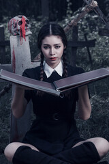 A young woman dressed in black, holding a grim reaper's scythe and a black magic wand, stands in the middle of a graveyard surrounded by graves, smoke, and human skulls. Halloween costume idea.