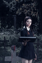 A young woman dressed in black, holding a grim reaper's scythe and a black magic wand, stands in the middle of a graveyard surrounded by graves, smoke, and human skulls. Halloween costume idea.