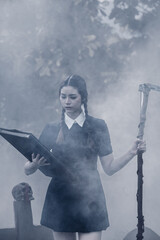 A young woman dressed in black, holding a grim reaper's scythe and a black magic wand, stands in the middle of a graveyard surrounded by graves, smoke, and human skulls. Halloween costume idea.