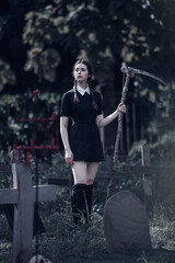 A young woman dressed in black, holding a grim reaper's scythe and a black magic wand, stands in the middle of a graveyard surrounded by graves, smoke, and human skulls. Halloween costume idea.