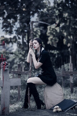 A young woman dressed in black, holding a grim reaper's scythe and a black magic wand, stands in the middle of a graveyard surrounded by graves, smoke, and human skulls. Halloween costume idea.