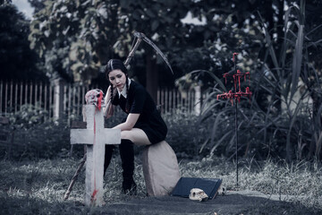 A young woman dressed in black, holding a grim reaper's scythe and a black magic wand, stands in the middle of a graveyard surrounded by graves, smoke, and human skulls. Halloween costume idea.