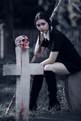A young woman dressed in black, holding a grim reaper's scythe and a black magic wand, stands in the middle of a graveyard surrounded by graves, smoke, and human skulls. Halloween costume idea.