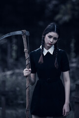 A young woman dressed in black, holding a grim reaper's scythe and a black magic wand, stands in the middle of a graveyard surrounded by graves, smoke, and human skulls. Halloween costume idea.