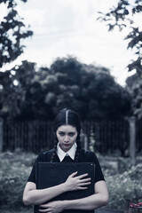 A young woman dressed in black, holding a grim reaper's scythe and a black magic wand, stands in the middle of a graveyard surrounded by graves, smoke, and human skulls. Halloween costume idea.