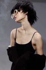 Fototapeta premium Stylish young woman with short hair wearing trendy round glasses and a black outfit against smoky dark background