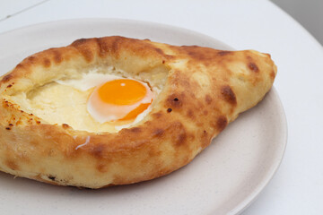 Food photo close up. Fresh tasty Adjarian style khachapuri with yellow yolk of an egg and sulguni cheese on white round plate on Georgian cafe table. Concept of eat, national cuisine, menu, lunch