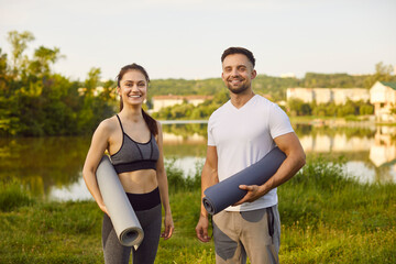 Smiling young man and woman holding yoga mats and looking at camera. Cheerful sporty couple standing on green grass near lake ready for outdoor workout or yoga session and enjoying active lifestyle.