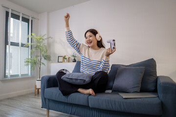 A young woman sits on a sofa wearing headphones while holding a game controller and playing a video game on her laptop. Her cheerful expression shows enjoyment and excitement.