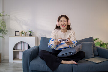 A young woman sits on a sofa wearing headphones while holding a game controller and playing a video game on her laptop. Her cheerful expression shows enjoyment and excitement.