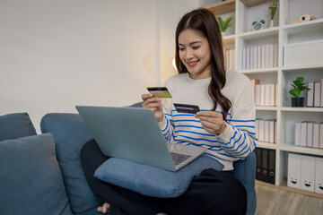 A young woman sits on a sofa using a laptop while holding credit cards, representing secure online payment and modern lifestyle. She smiles confidently while shopping from home.