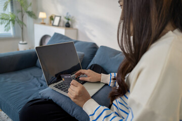 A young woman sits on a sofa using a laptop while holding credit cards, representing secure online payment and modern lifestyle. She smiles confidently while shopping from home.