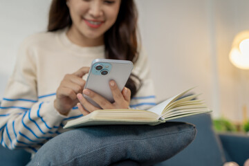 A young woman sitting comfortably on a sofa holding a smartphone while smiling and reading a message, with an open notebook resting on her lap in a bright cozy room.
