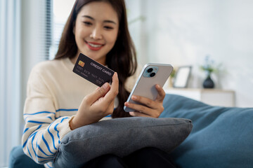 A young woman is sitting on a sofa holding a smartphone and credit card, smiling as she makes an online transaction. She looks relaxed and confident while shopping at home.
