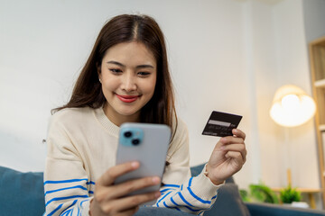A young woman is sitting on a sofa holding a smartphone and credit card, smiling as she makes an online transaction. She looks relaxed and confident while shopping at home.