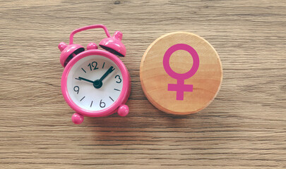 Pink Alarm Clock Next to Female Venus Symbol on Wood Block Representing Women's Biological Clock and Fertility