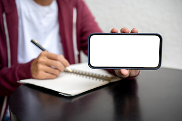 Man student showing blank screen smartphone in horizontal while writing in notebook on table in cafe