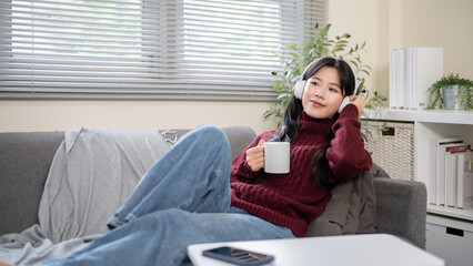 Asian woman with headphone holding coffee sitting leaning on sofa with phone on table in living room