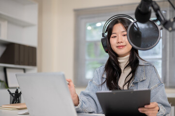 Woman wears mic headset holding clipboard and hosting radio or podcast with laptop at home or studio