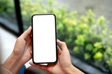 Close up of a woman hands holding white screen smartphone with blurred garden outside a glass wall.