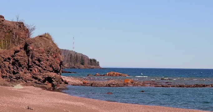 Waves roll onto a beach at the rocky shore of Lake Superior, as seagulls fly over and float on the blue lake water, on a sunny day in Minnesota.