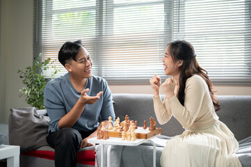 Asian woman smiling after winning a chess match on wooden board playing with a man at sofa table.