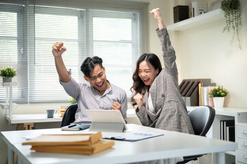 Asian businessman and woman coworker looking at tablet raising a fist in excited at table in office