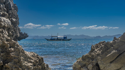 The blue ocean is visible through the gap between the coastal cliffs. A traditional Filipino bangka boat floats in the sea. Mountains on the horizon. Azure sky, clouds. Philippines. Palawan.