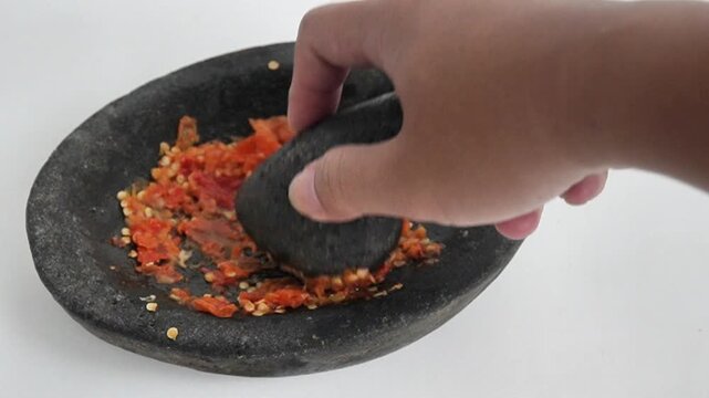 A close-up shot of a hand grinding red chili peppers and seeds with a traditional stone pestle in a mortar (cobek and ulekan) to make spicy Indonesian sambal. Onion sauce, sambal bawang