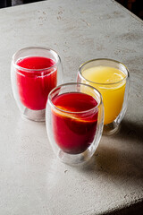 Bright red and orange juices sit elegantly in clear glass cups on a neutral surface