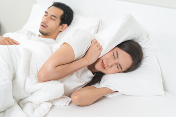 Snoring couple health asian man snore and sleeping at home while woman insomnia annoyed, bad noise cover ears with pillow, sleep problem apnea and relationship, wife annoyed husband snoring in bed.